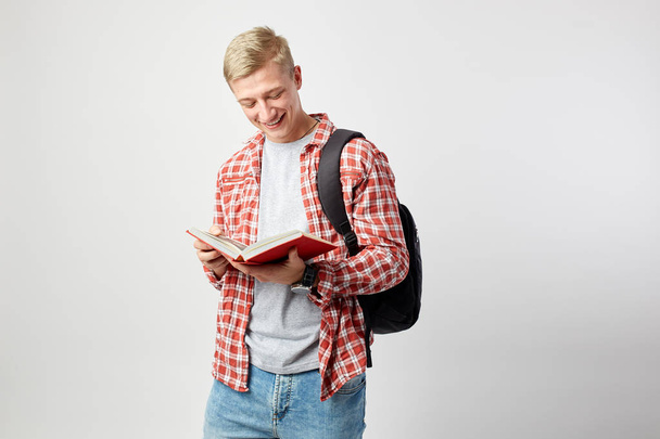 Young blond guy with black backpack on his shoulder dressed in a white t-shirt, red checkered shirt and jeans reads a book on the white background  in the studio - Photo, Image