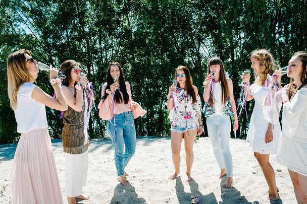 Счастливы стильные сексуальные девушки с цветочными бутоньерками на руках, пьющие шампанское из бокалов на песчаном пляже. Вечеринка в стиле бохо. Девичий вечер девичников
  - Фото, изображение