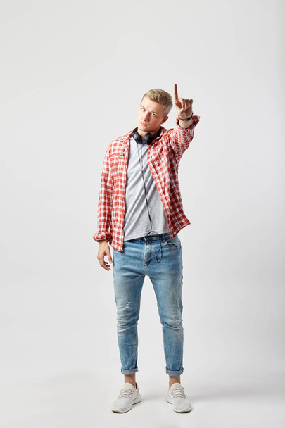 Blond guy with headphones on his neck dressed in a white t-shirt, red checkered shirt and jeans stands on the white background in the studio and point up with his finger - Photo, image