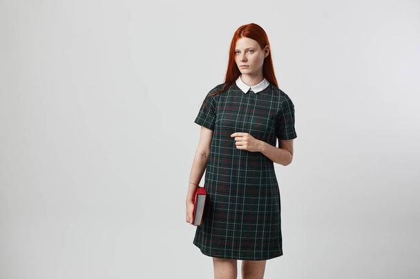 Charming redhead long-haired girl in a green checkered dress holds a book in her hand on the white background in the studio - Photo, Image
