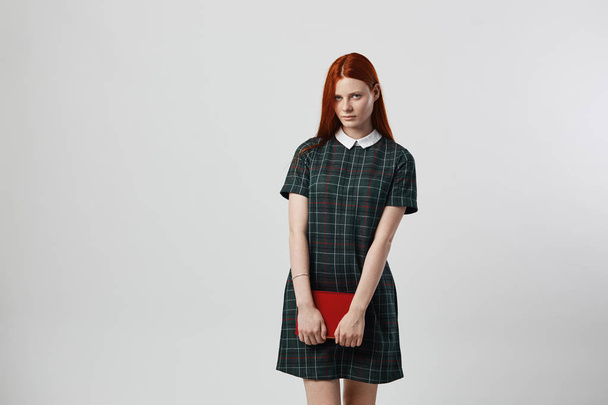Charming redhead long-haired girl in a green checkered dress holds a book in her hands on the white background in the studio - Φωτογραφία, εικόνα