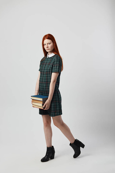Charming redhead long-haired girl in a green checkered dress walks with books in her hands on the white background in the studio - Photo, Image
