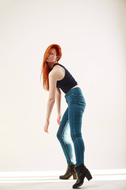 Charming redhead long-haired girl dressed in black top and jeans is posing on the white background in the studio - Foto, afbeelding