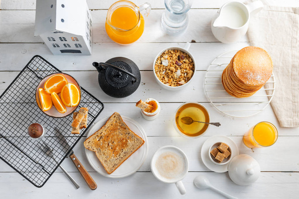 Healthy breakfast eating concept, various morning food - pancakes, soft-boiled egg, toast, oatmeal, granola, fruit, coffee, tea, orange juice, milk on white wooden table - Foto, Imagem