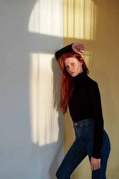 Beautiful redhead long-haired girl dressed in black top and jeans is posing against the wall with a reflection of the sunny window - Фото, изображение