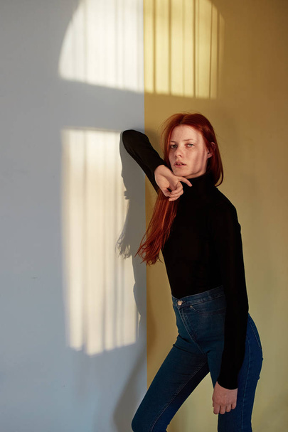 Beautiful redhead long-haired girl dressed in black top and jeans is posing against the wall with a reflection of the sunny window - Foto, afbeelding