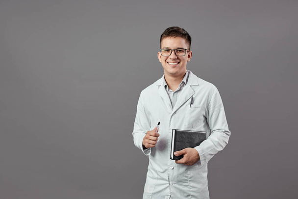 Intelligent smiling guy in glasses dressed in a white coat is holding a notebook and a pan on a gray background - Foto, Imagem
