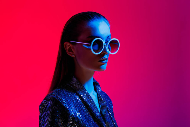 Fashion girl with long hair and round sunglasses in a black shining dress poses in neon light in the studio - Фото, зображення
