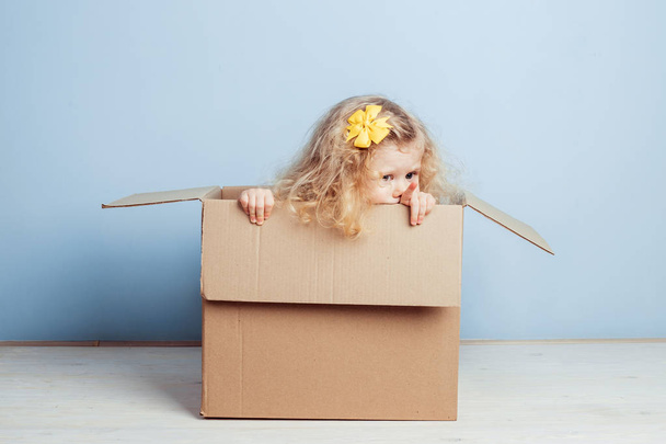 Little curly girl with yellow flower on her hair sits in the cardboard box on the background of blue wall. - Foto, imagen