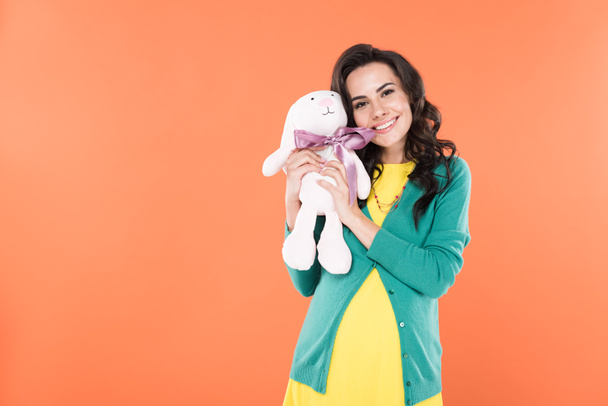 Charming brunette pregnant woman holding toy rabbit and smiling isolated on orange  - Foto, afbeelding