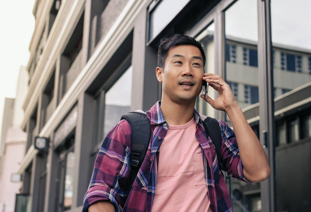 Close up of young Asian man in a plaid shirt walking along a city sidewalk, talking on a cellphone - Foto, Imagen