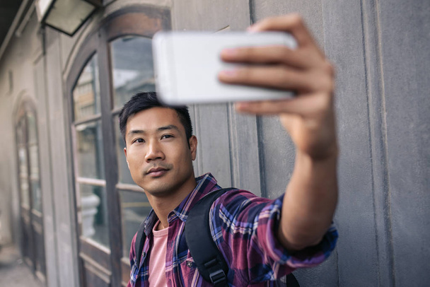 Confident young Asian man standing on a sidewalk in the city taking a selfie with his cellphone - Photo, Image