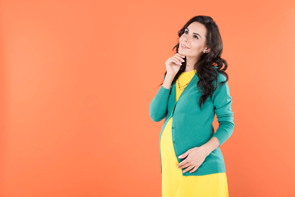 Pensive pregnant woman with curly hair looking up with smile isolated on orange - Photo, Image