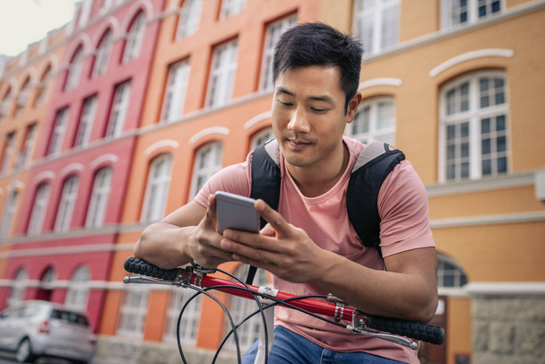 Young Asian man sitting on his bike in front of colorful buildings in the city sending a text on his cellphone - Foto, Imagen