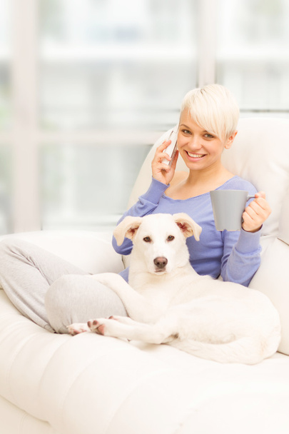 kadın ile köpek bir kupa holding ve telefon ederek kanepe - Fotoğraf, Görsel