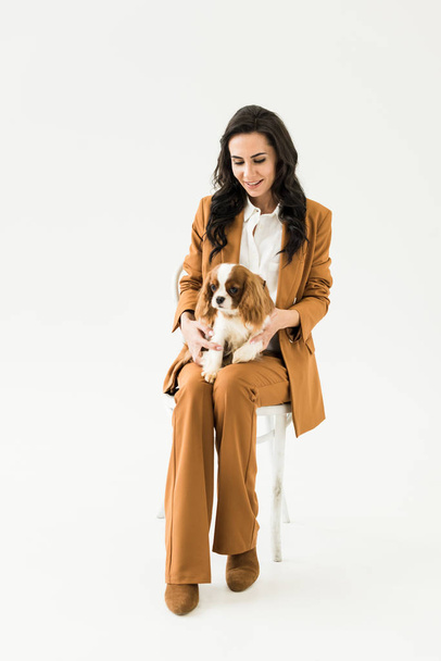 Smiling young woman in brown suit sitting on chair and holding dog on white background - 写真・画像