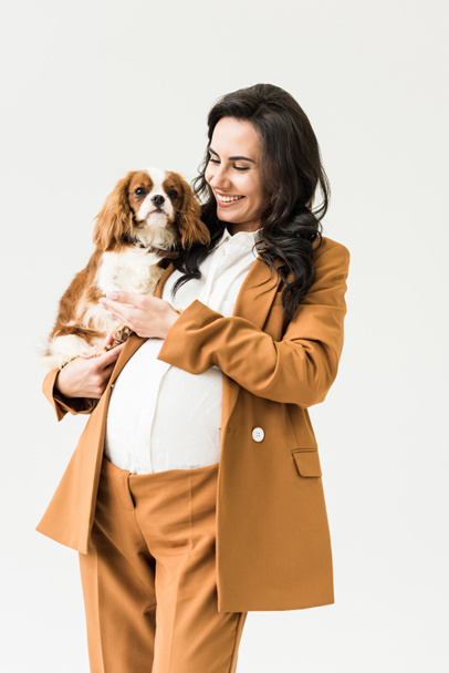 Joyful pregnant woman in suit holding dog isolated on white - 写真・画像