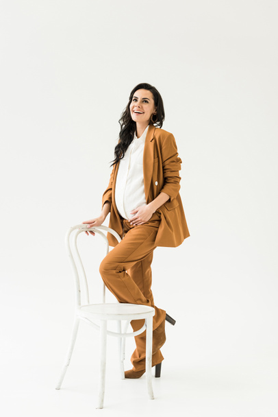 Laughing pregnant woman in brown suit posing with chair on white background - Zdjęcie, obraz