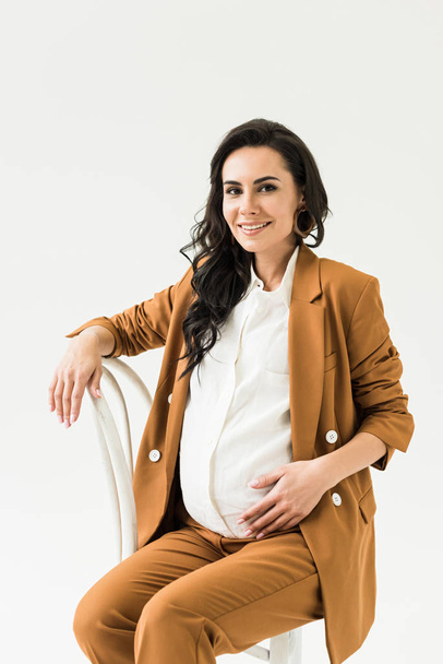 Smiling brunette pregnant woman sitting on chair isolated on white  - Foto, Imagem
