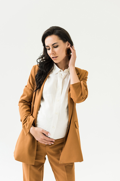 Pensive brunette pregnant woman in brown suit touching belly isolated on white - Photo, Image