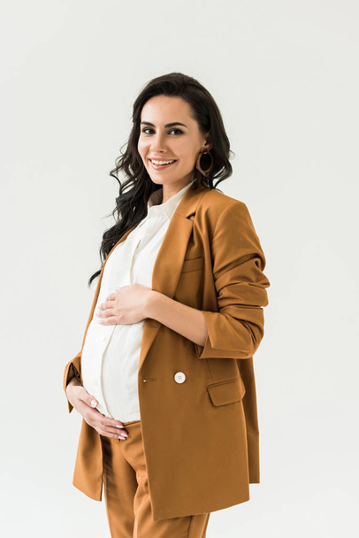 Blissful pregnant woman in brown suit touching tummy and looking at camera isolated on white - Fotografie, Obrázek