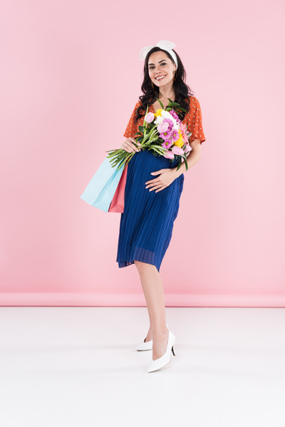 Pregnant woman in blue skirt holding flowers and shopping bags on pink background - Photo, Image