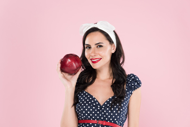 Stylish brunette woman in polka-dot dress holding ripe red apple isolated on pink - Photo, Image