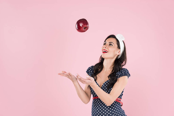 Smiling pregnant girl in dress throwing red apple isolated on pink - Fotó, kép