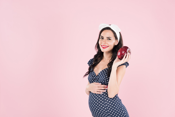 Όμορφη έγκυος γυναίκα με πουά φόρεμα εκμετάλλευση κόκκινο μήλο που απομονώνονται σε ροζ - Φωτογραφία, εικόνα