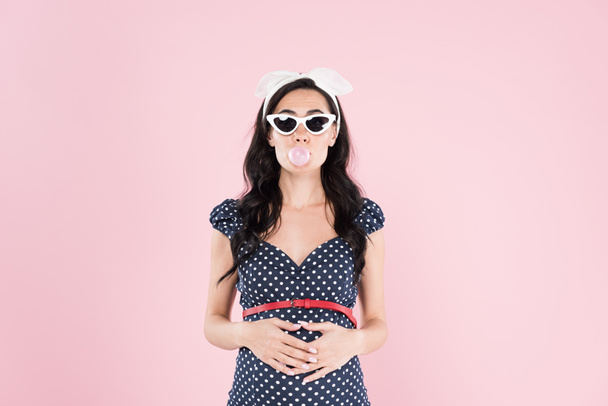 Brunette pregnant woman chewing bubble gum and touching belly isolated on pink - Photo, Image