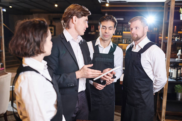 Serious confident restaurant manager  using digital tablet and pointing at screen while discussing seating chart with waiters before banquet - Photo, Image