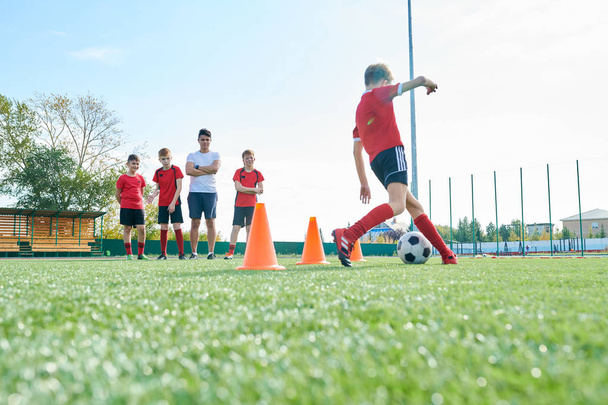 Full length portrait of junior football team training outdoors in sunlight with focus on  unrecognizable boy leading ball between orange cones, copy space - Zdjęcie, obraz
