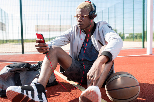 Portrait d'un Africain contemporain assis sur le sol du terrain de basket et écoutant de la musique via smartphone, espace de copie
 - Photo, image