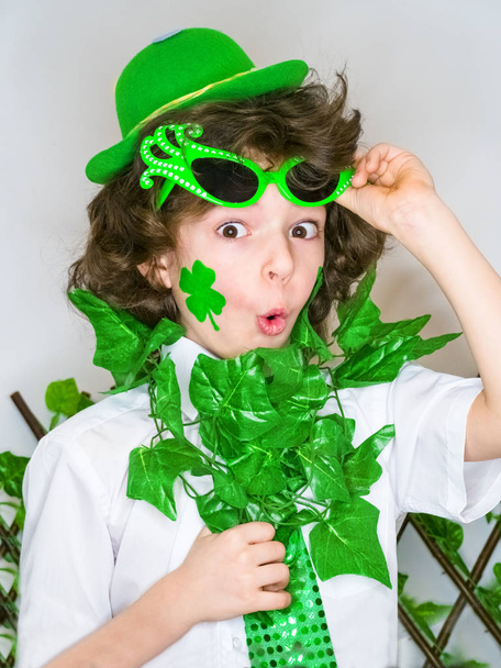 Funny St Patricks day little curly boy wearing a green hat and green carnival accessories shocked with an open mouth. light background - Foto, afbeelding