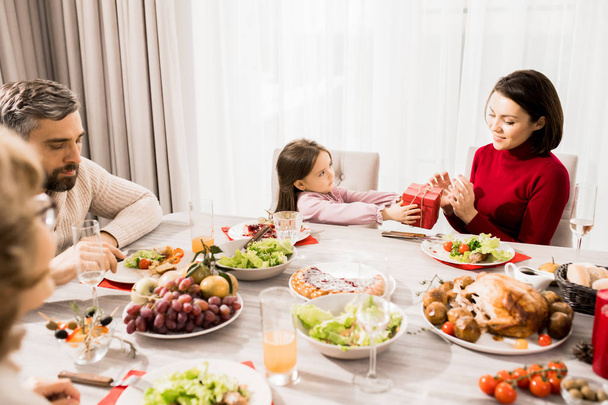 Warm-toned portrait of big happy family enjoying Christmas dinner together, focus on little girl giving present to mom - Foto, immagini