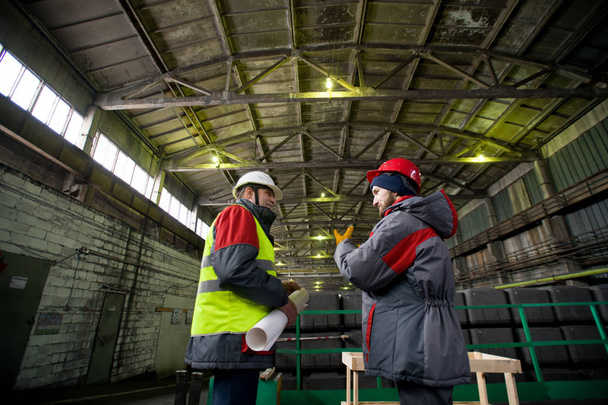 Retrato retrovisor de dos trabajadores industriales que usan chaquetas y sombreros calientes discutiendo la producción de pie en el taller
 - Foto, Imagen