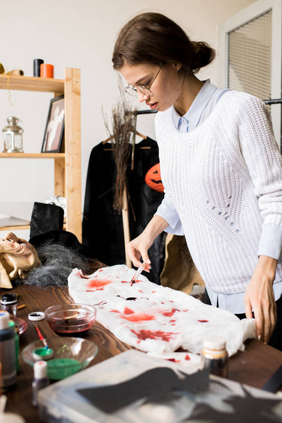 Poważny skoncentrowany projektant odzieży w sweter stojący na stole i upuszczając farby z krwi kolor na gazę podczas robienia Halloween kostium w warsztacie - Zdjęcie, obraz