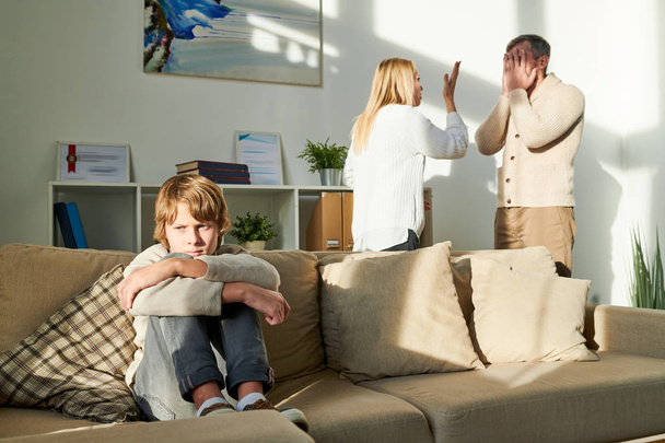 Расстроенный депрессивный подросток, сидящий на диване и обнимающий колени, страдающий от конфликта родителей, мать и отец ссорятся на заднем плане
 - Фото, изображение