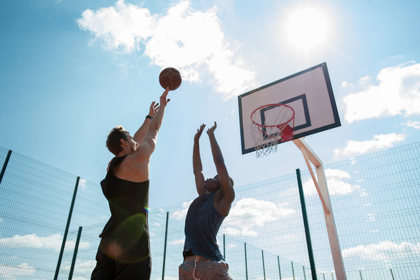 Vista de ángulo bajo de dos hombres jugando baloncesto y saltando alcanzando la pelota por aro contra el cielo azul iluminado por el sol, espacio de copia
 - Foto, Imagen