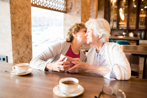 Tartalom érett hölgy kardigán ül az asztalnál, és megcsókolta barátja, miközben ajándék neki a modern kávézóban - Fotó, kép