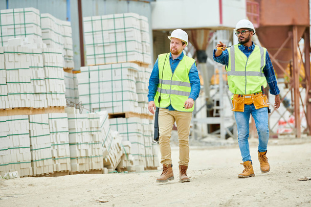Zwei junge Männer in Bollenhut und Weste zeigen bei einer Baustellenkontrolle auf Distanz, während sie in der Nähe von Baustoffstapeln spazieren gehen - Foto, Bild