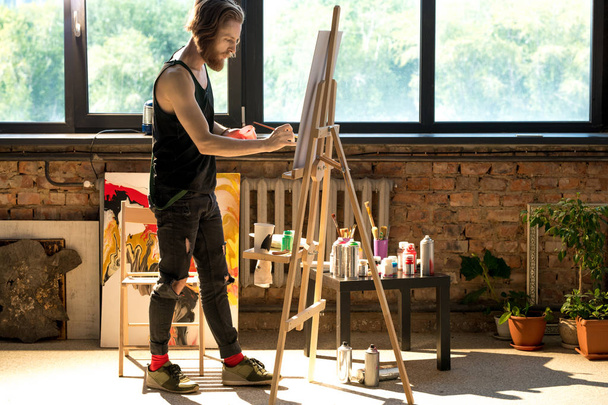 Retrato de comprimento total de belo artista masculino pintura no cavalete enquanto desfruta de trabalho em estúdio de arte iluminado pela luz solar, espaço de cópia
 - Foto, Imagem