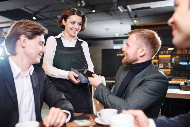 Glimlachend mooi jong serveerster in zwarte schort geven Terminal voor betaling aan zakenman in modern restaurant - Foto, afbeelding
