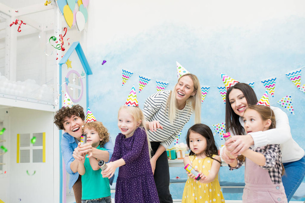 Wesołe ekstatyczne młode matki i dzieci w kolorowe kapelusze party stoi w niebieskim pokoju gier i pęka krakersy podczas zabawy na imprezie - Zdjęcie, obraz