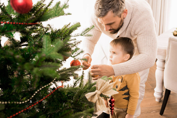 warmfarbiges Porträt eines glücklichen Vaters, der mit seinem entzückenden kleinen Sohn den Weihnachtsbaum schmückt, Kopierraum - Foto, Bild