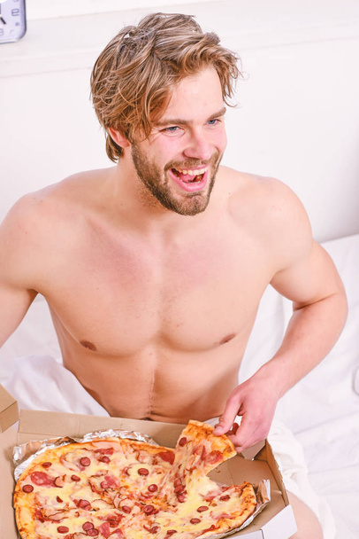 Shirtless handsome young man with pizza on bed. Guy holds pizza box sit bed in bedroom or hotel room. Portrait of lazy muscular man eating pizza while laying on a bed at home. Pizza on bed. - Fotografie, Obrázek