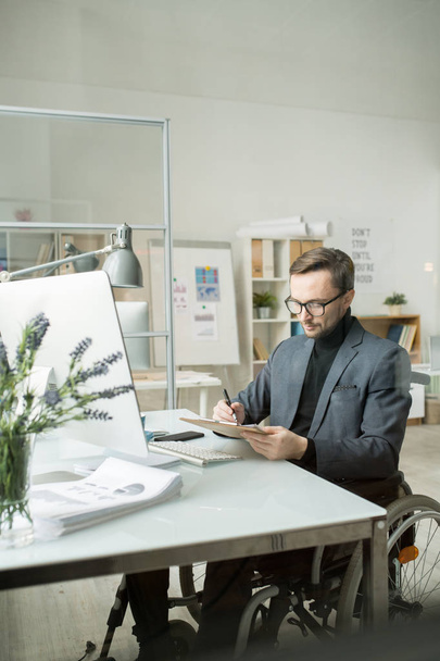 Vakava vammainen silmälasipäinen liikemies, joka tekee paperitöitä työpaikallaan nykyaikaisen toimiston tietokoneen edessä
 - Valokuva, kuva