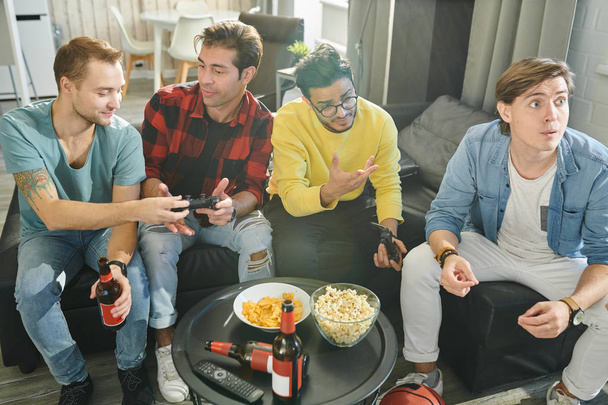 Los hombres jóvenes descansando en el sofá con palomitas de maíz y bebidas alcohólicas, que juegan videojuegos a su vez
 - Foto, imagen