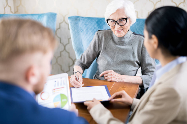 Mujer mayor sonriente con anteojos firmando un contrato en la mesa con agentes de seguros
 - Foto, Imagen