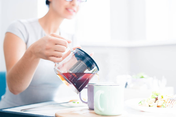 Closeup of unrecognizable young woman pouring black tea from glass kettle while enjoying lunch in sunlit cafe, copy space - Foto, Imagem